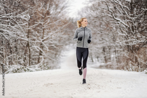 Full length of happy middle-aged sportswoman running in nature at snowy winter day. Outdoor fitness, cardio exercises, exercises in nature