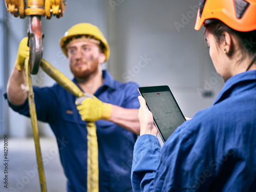 Industrial worker fixing hoist sling on indoor crane, female colleague controlling with digital tablet photo