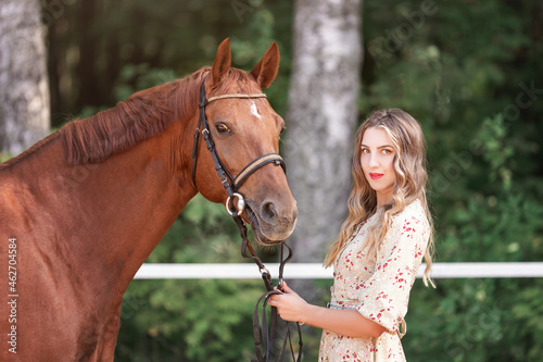 Beautiful young woman in elegant lifestyle dress standing near her pet red horse at farm at summer nature