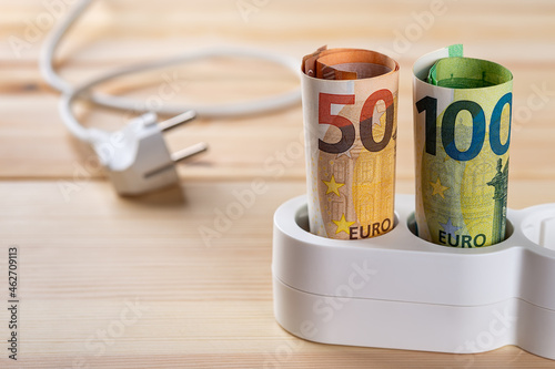 Euro money banknotes inserted into a white extension socket on a wooden surface. Expensive energy and rise in electricity prices concepts. Increasing of electricity cost.