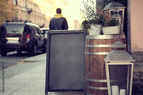 An advertising black wooden sign on the street in front of a cafe, bar, diner, surrounded by flower pots and stylish lamps. A place for a text, poster, or public information.