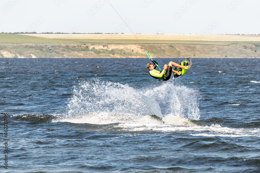 Obraz premium Young adult caucasian fit male person enjoy riding kite surf board making extreme trick stunt bright sunny day against blue sky at sea or ocean shore. Watersport adrenaline fun adventure acitivity