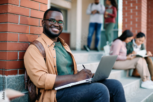 Happy black university student writes notes while using laptop outdoors and looking at camera.