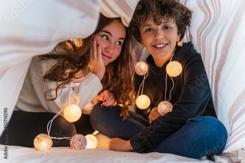 Portrait of happy brother and sister with chain of lights underneath bedcover photo