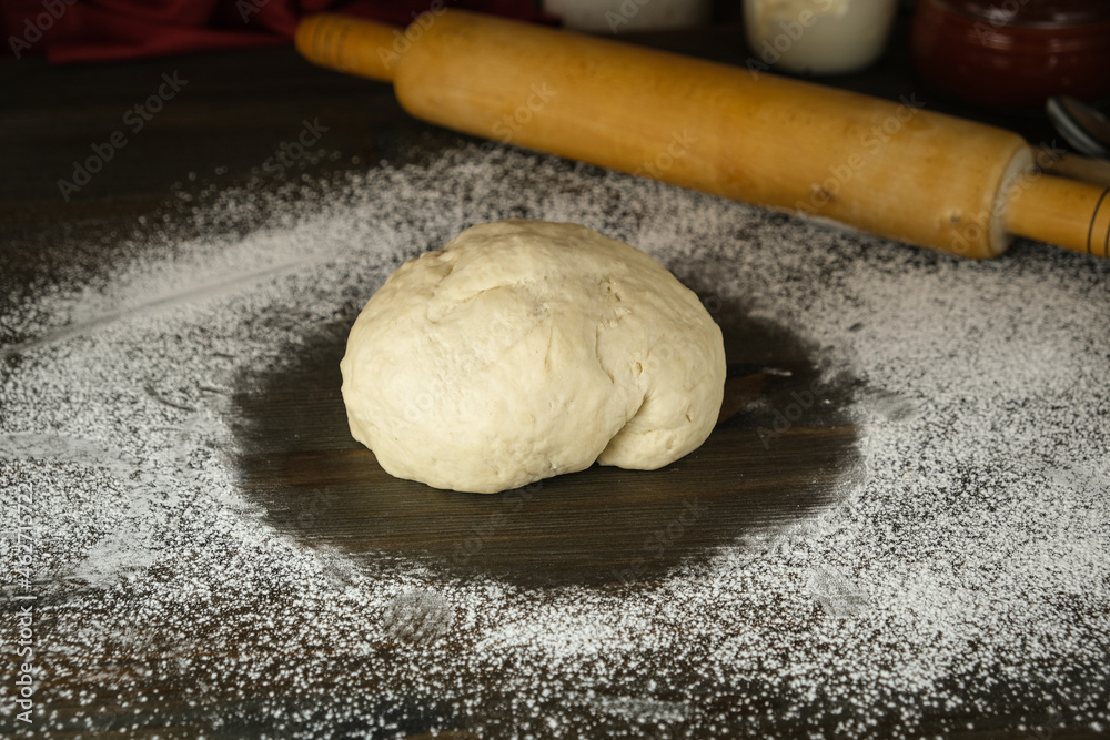 a mixture of yeast dough lies on a dark board sprinkled with white flour. a wooden rolling pin is located behind the dough. the process of making homemade pies and pizza. bread sourdough. a mixture of yeast dough lies on a dark board sprinkled with white flour. a wooden rolling pin is located behind the dough. the process of making homemade pies and pizza. bread sourdough.