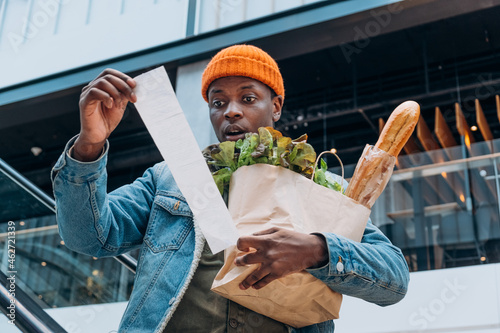 Doubting African-American person in denim jacket looks at sales paper receipt total holding pack with food products on escalator photo