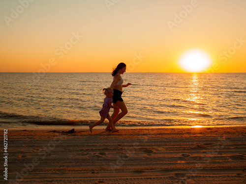 A young woman and a little girl run along the sandy sea beach along the water. In the background, the evening sun sets over the horizon.