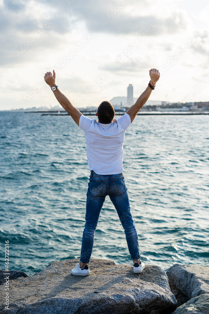 Spain, Barcelona, back view of man with raised arms standing in front of the sea Spain, Barcelona, back view of man with raised arms standing in front of the sea