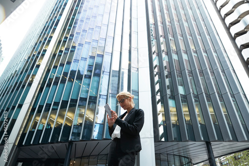 Blond businesswoman using phone while standing against modern office building in financial district of city photo