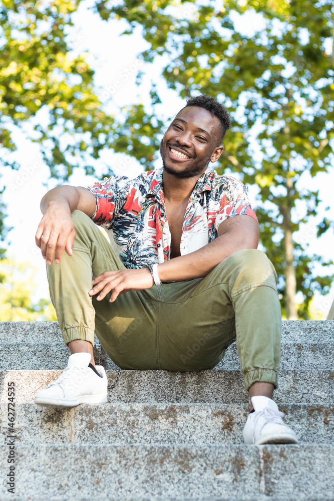 Smiling young man in casuals sitting on steps against trees Smiling young man in casuals sitting on steps against trees