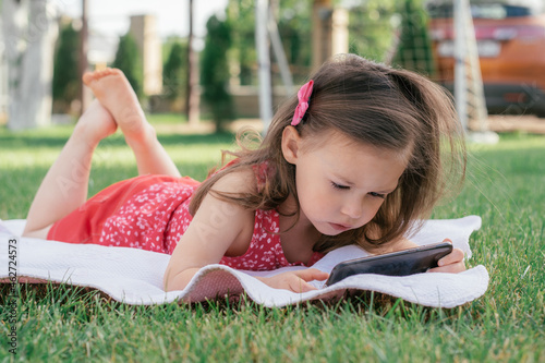 Little 3-4 girl in red clothes lies on blanket on green grass and looks into mobile phone. Children, using gadgets