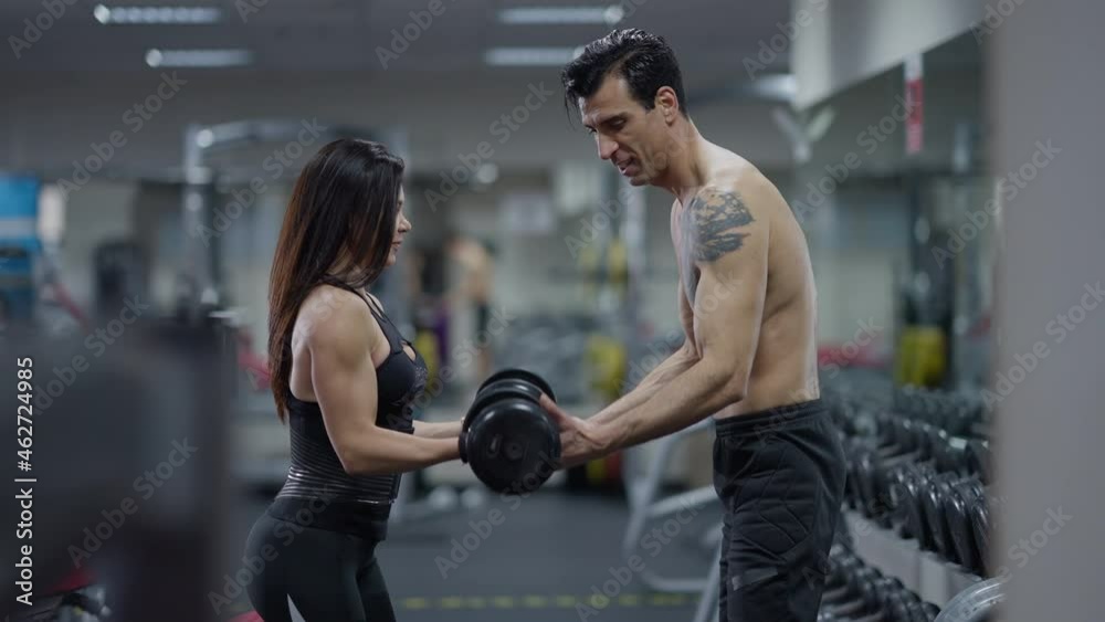 Side view smiling fit Middle Eastern man training confident Caucasian young woman in gym. Motivated sportswoman lifting dumbbells as sportsman endorsing supporting client. Bodybuilding and fitness Side view smiling fit Middle Eastern man training confident Caucasian young woman in gym. Motivated sportswoman lifting dumbbells as sportsman endorsing supporting client. Bodybuilding and fitness