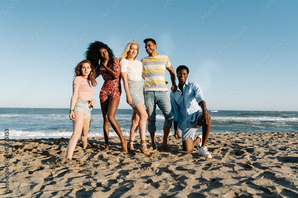 Young friends standing on sand against water during sunny day Young friends standing on sand against water during sunny day
