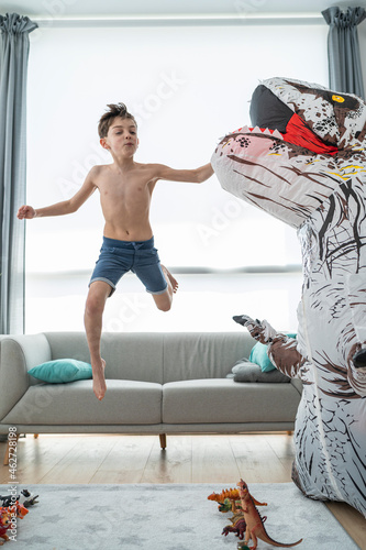 Shirtless boy jumping from sofa while fighting with large toy dinosaur photo