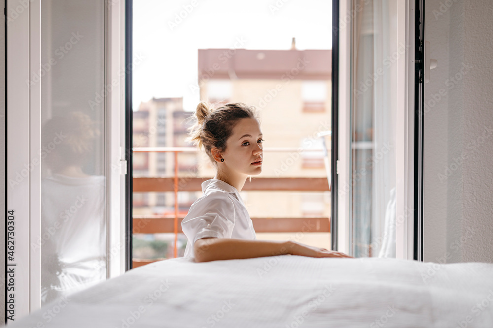 Side view of thoughtful young woman looking away while sitting by bed at home