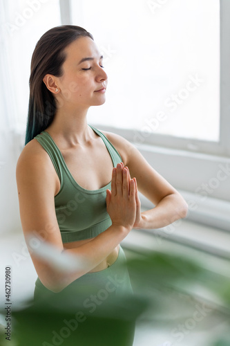 Beautiful young woman meditating while standing by window. Palms together. Blurred foreground.