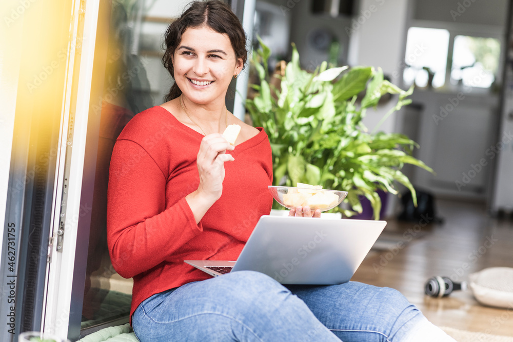 Smiling young woman eating fruit while using laptop at home Smiling young woman eating fruit while using laptop at home