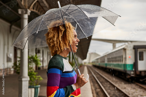 Side view of cheerful young woman enjoying while standing with umbrella at railroad station during monsoon