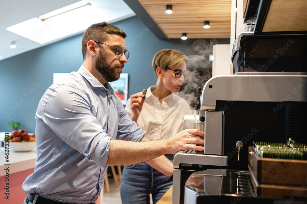 Bearded businessman making coffee while standing by female colleague at office cafeteria during break Bearded businessman making coffee while standing by female colleague at office cafeteria during break