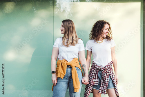 Smiling beautiful women holding hands while looking away against wall photo