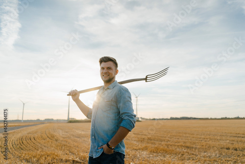 Portrait of smiling young man holding pitchfork standing on stubble field photo