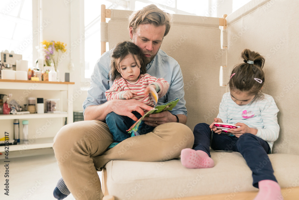 Father reading book to daughter with her sister playing Father reading book to daughter with her sister playing