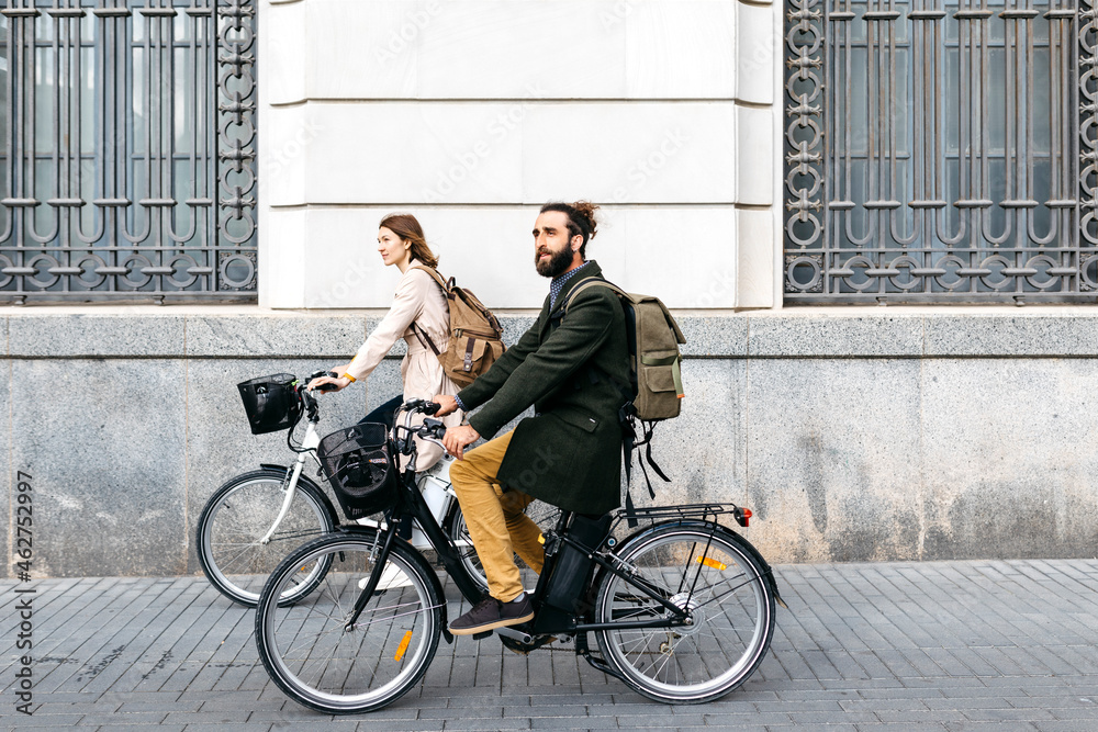 Couple riding e-bikes in the city passing a building Couple riding e-bikes in the city passing a building