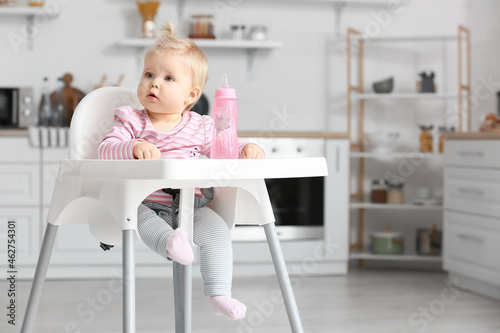 Cute baby girl with bottle of water sitting on high chair in kitchen