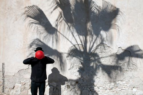 Morocco, Essaouira, man wearing a bowler hat holding red balloon in front of his face at a wall photo
