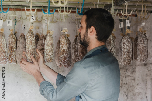 Young bearded man examining salami sausages hanging at smokehouse photo