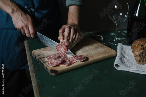 Hands of man cutting salami slices on board at table photo