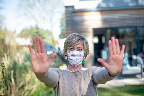 Portrait of mature woman wearing protective face mask while standing with stop gesture against house during epidemic lockdown photo