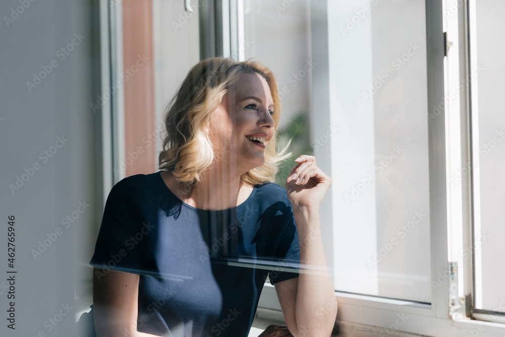 Happy blond woman in sunlight looking out of window Happy blond woman in sunlight looking out of window