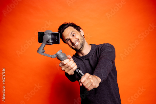 Cheerful man taking selfie while holding gimbal with smart phone against orange background photo