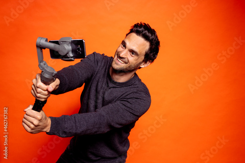 Playful handsome man taking selfie through smart phone on gimbal against orange background photo