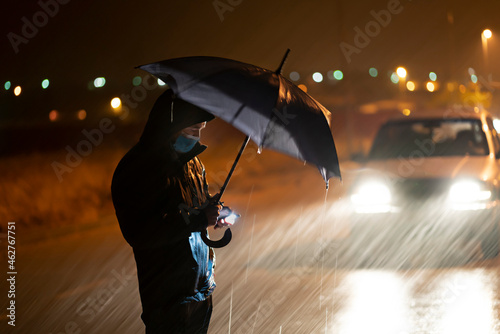 Young man with umbrella and face mask using mobile phone while standing against breakdown car during rainfall photo