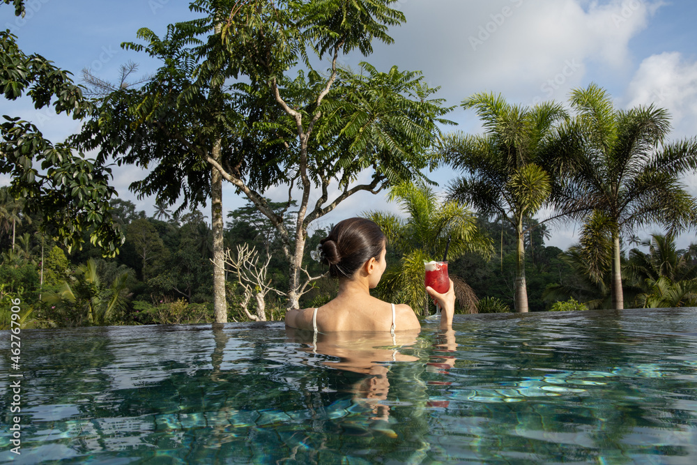 Fototapeta premium Woman drinking cocktail on infinity pool with stunning tropical view. Back view