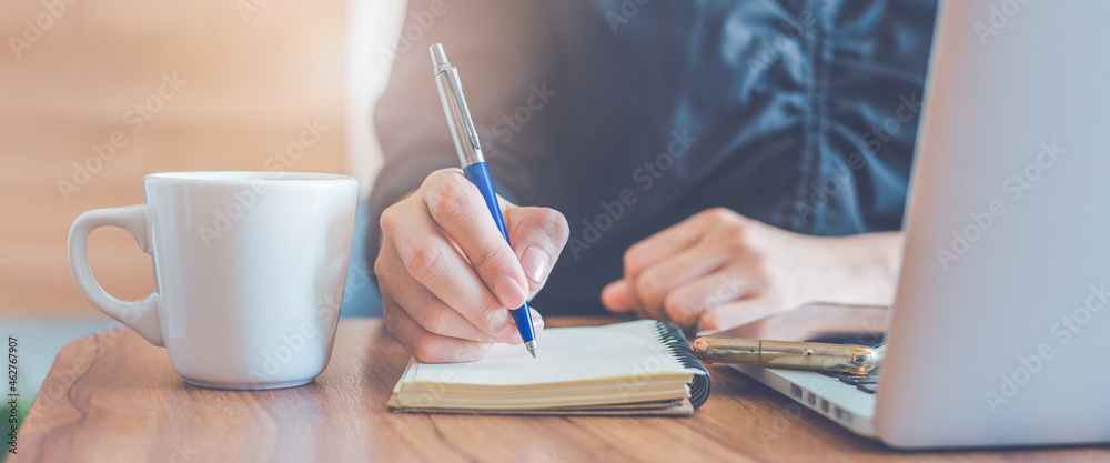 Business woman hand writing on a notepad with a pen and use a laptop computer in the office.