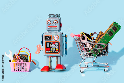 Studio shot of vintage robot toy standing between miniature shopping cart and basket filled with electronic equipment photo