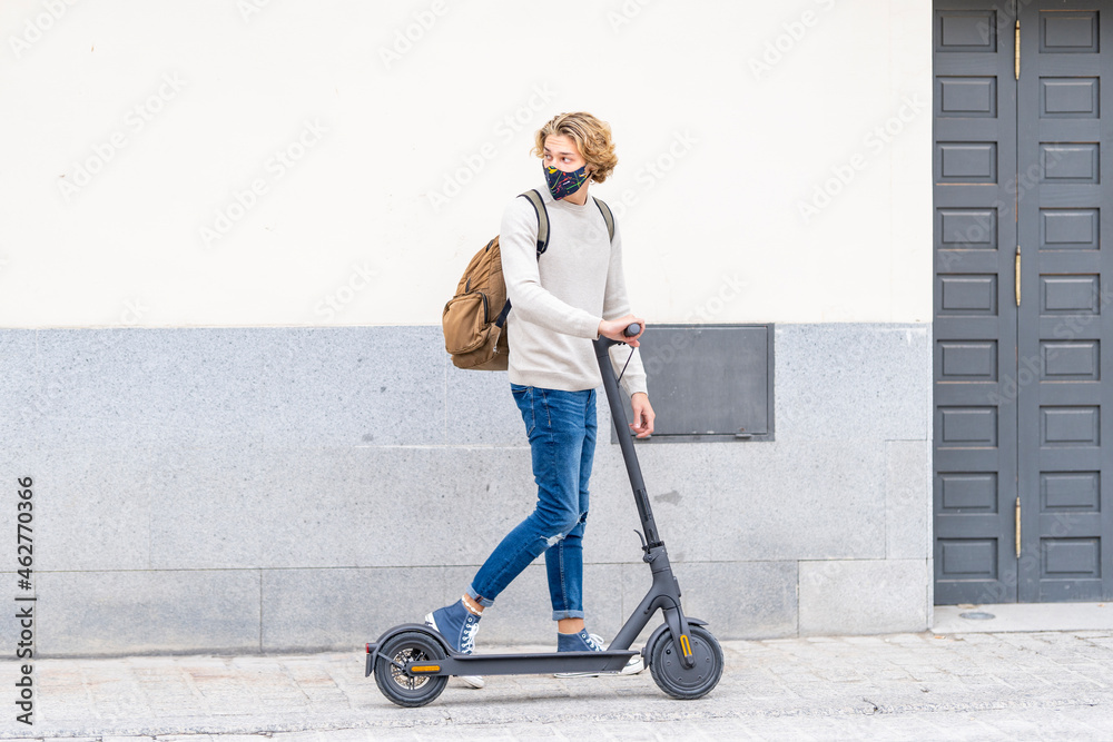 Young man wearing protective face mask standing with electric push scooter on footpath Young man wearing protective face mask standing with electric push scooter on footpath