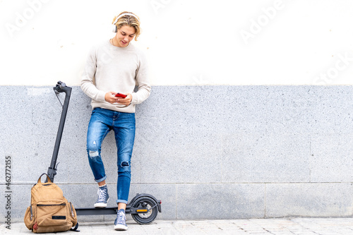 Man with backpack and electric push scooter using mobile phone while standing against wall
