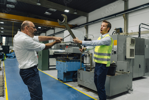 Businessman and man in reflective vest playfighting with large wrenches in a factory photo