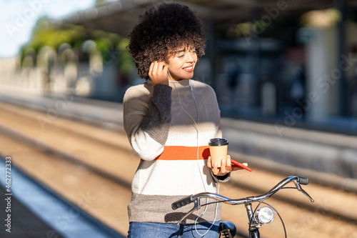 Smiling young woman with bicycle holding reusable cup while enjoying music at railroad station photo