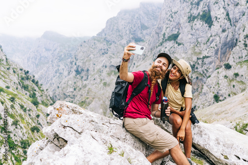 Smiling couple taking selfie while sitting on rock at Ruta Del Cares, Asturias, Spain