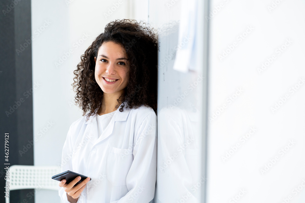Smiling female doctor with smart phone leaning by wall at clinic Smiling female doctor with smart phone leaning by wall at clinic