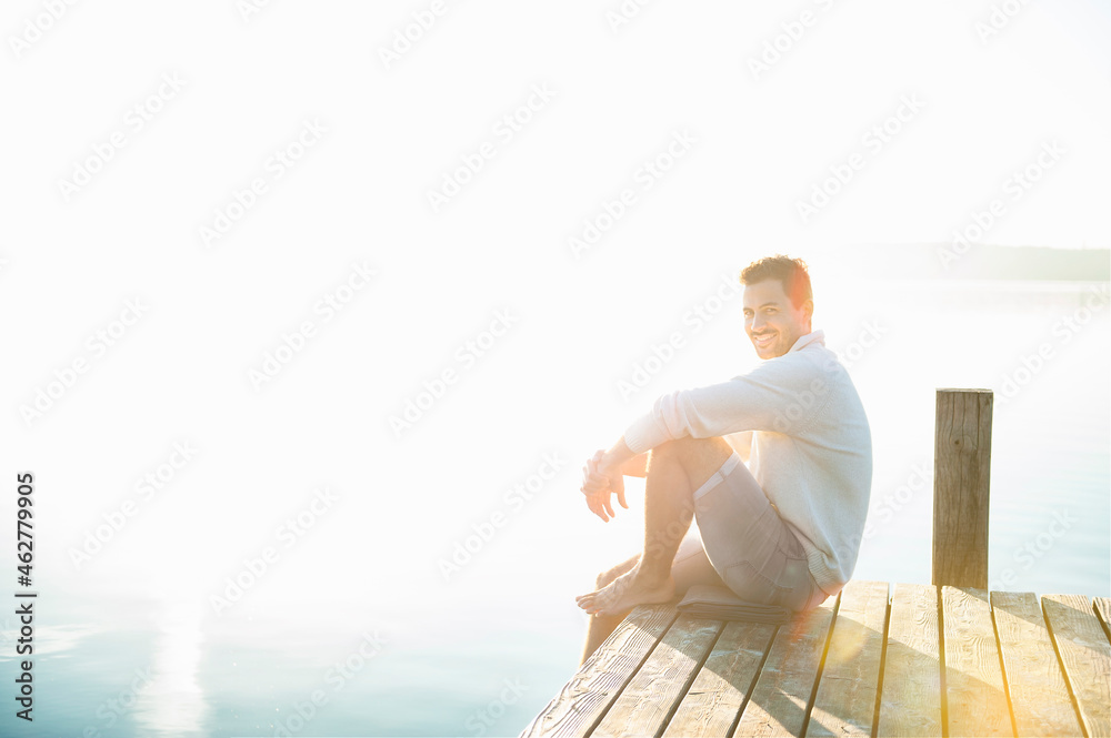 Portrait of smiling young man sitting on jetty at lake by sunrise Portrait of smiling young man sitting on jetty at lake by sunrise