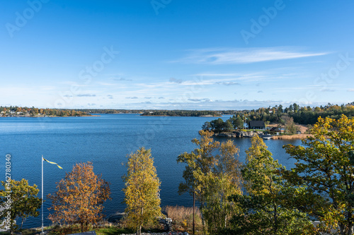 Swedish coast in autumn. Beautiful colorful panoramic view of the rocky shores of the Baltic sea bay with golden leaves trees and evergreens pines. Dark wooden small glass house with flag of Sweden.