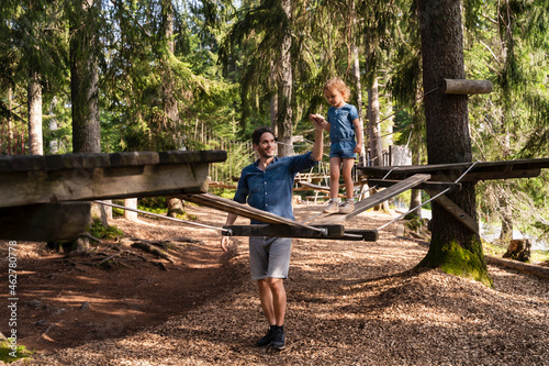 Father assisting little daughter walking across small suspension bridge on forest obstacle course