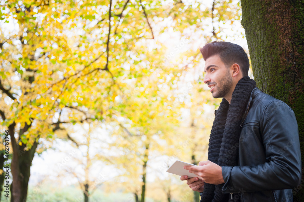 Handsome young man holding digital tablet while looking away and standing tree trunk at park Handsome young man holding digital tablet while looking away and standing tree trunk at park