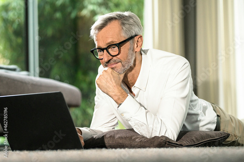 Smiling senior man with grey hair in modern design living room working on laptop photo